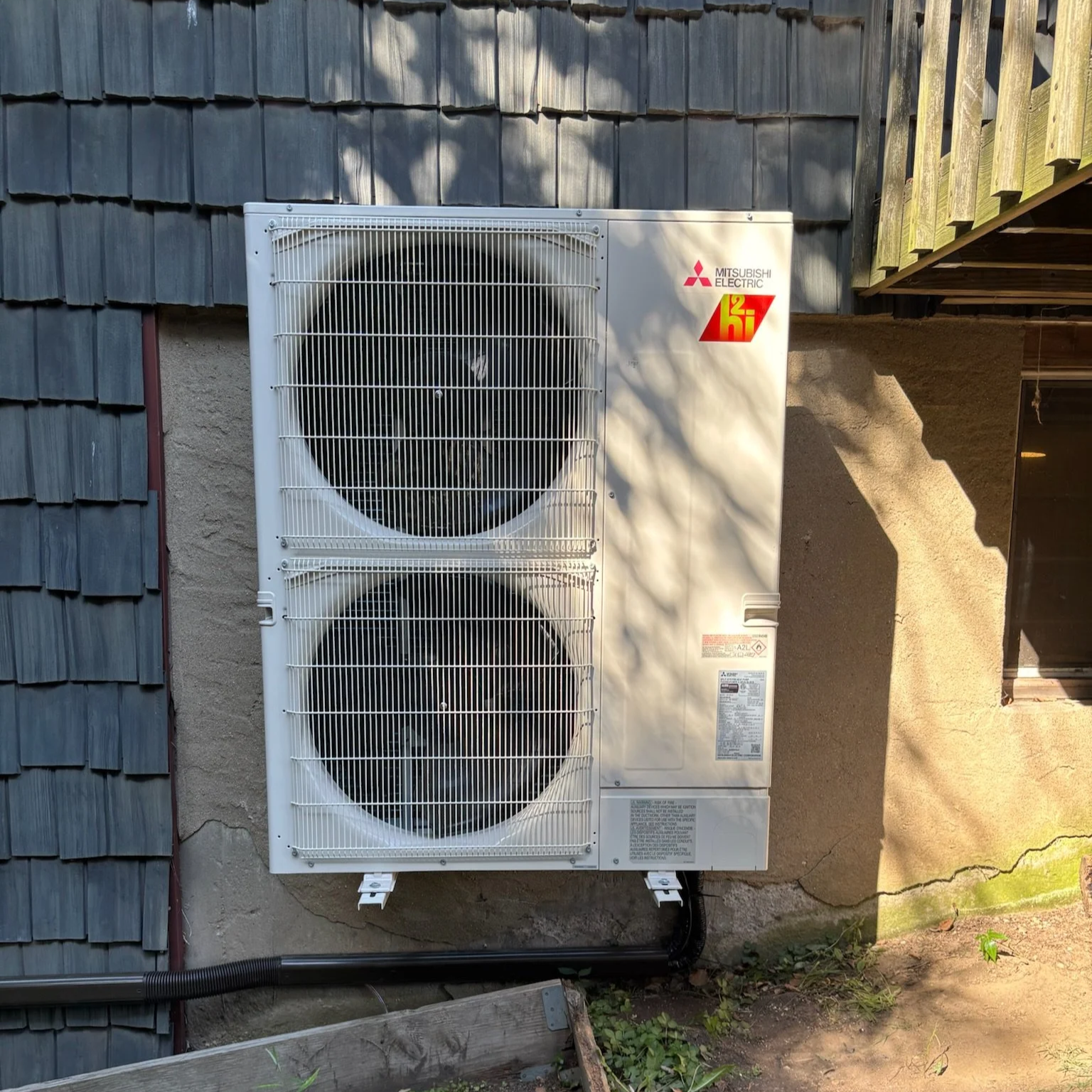 Outdoor Mitsubishi Electric air conditioning unit with two large fans behind protective metal grates, mounted on an exterior wall of a building, with shadows cast over it.