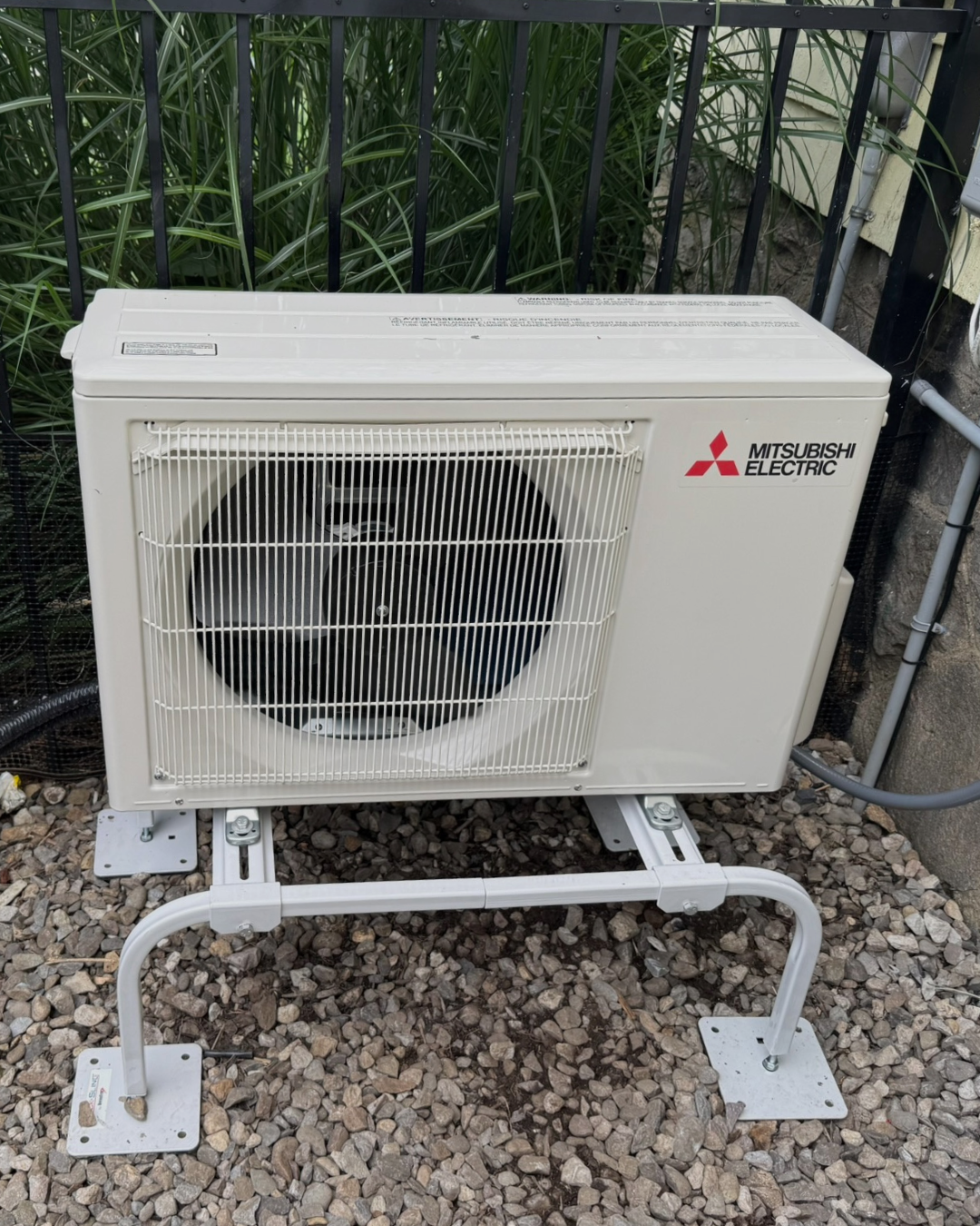 An outdoor Mitsubishi Electric air conditioning unit on a gravel surface with green plants and a fence in the background.