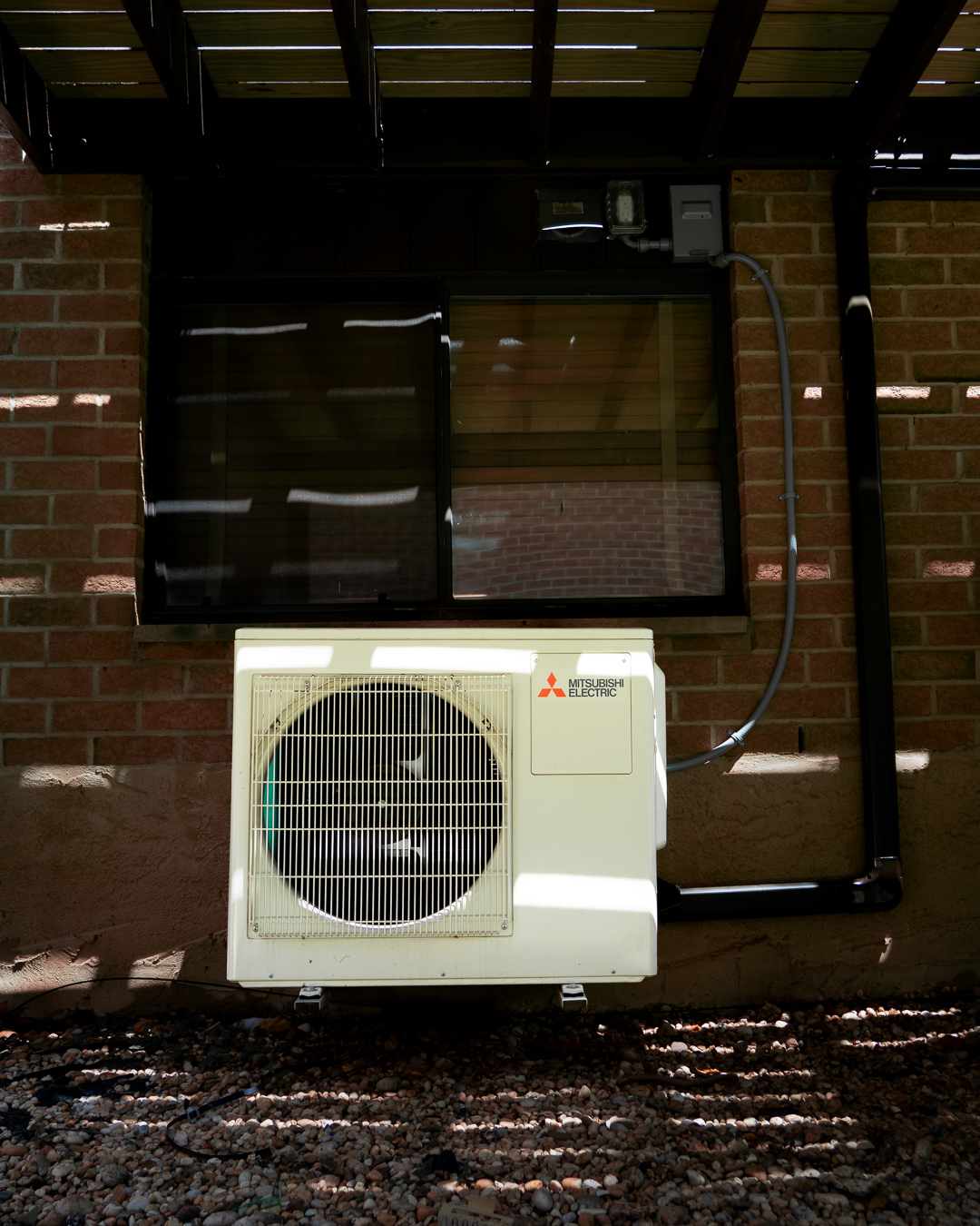 Exterior view of a brick building with an air conditioning unit mounted outside. The unit is a Mitsubishi Electric model, placed on the ground in front of a window with a black frame. Shadows and light create striped patterns on the wall and ground.