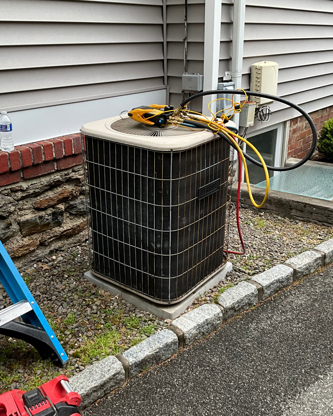 HVAC unit on a concrete pad outside a building with colored cables and tools on top, next to a brick and siding wall.