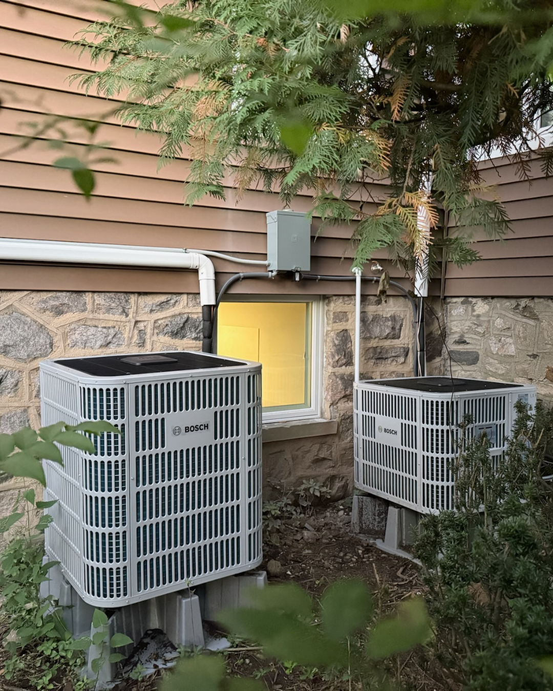 Two Bosch air conditioning units outside a house, surrounded by green plants and trees, with a small window in the stone wall behind them.