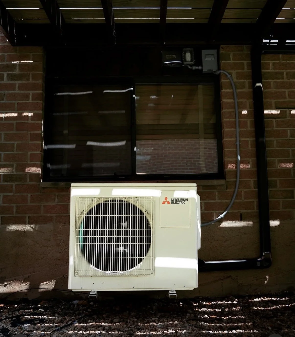 Exterior view of a brick building with a Mitsubishi Electric air conditioning condenser unit mounted on the wall, and a window with black screen and metal frame.