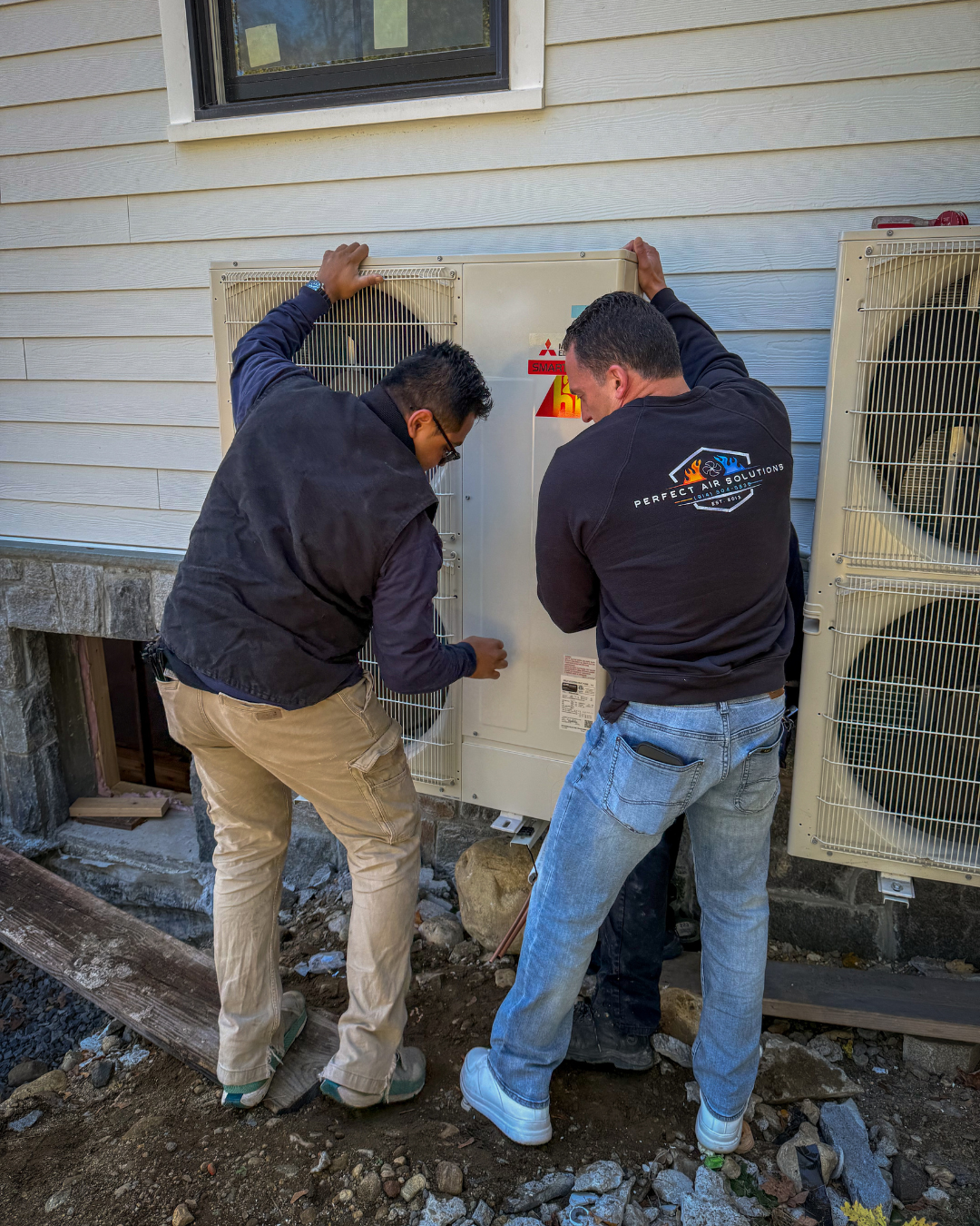 Two technicians working on an outdoor HVAC unit next to a house with vinyl siding.
