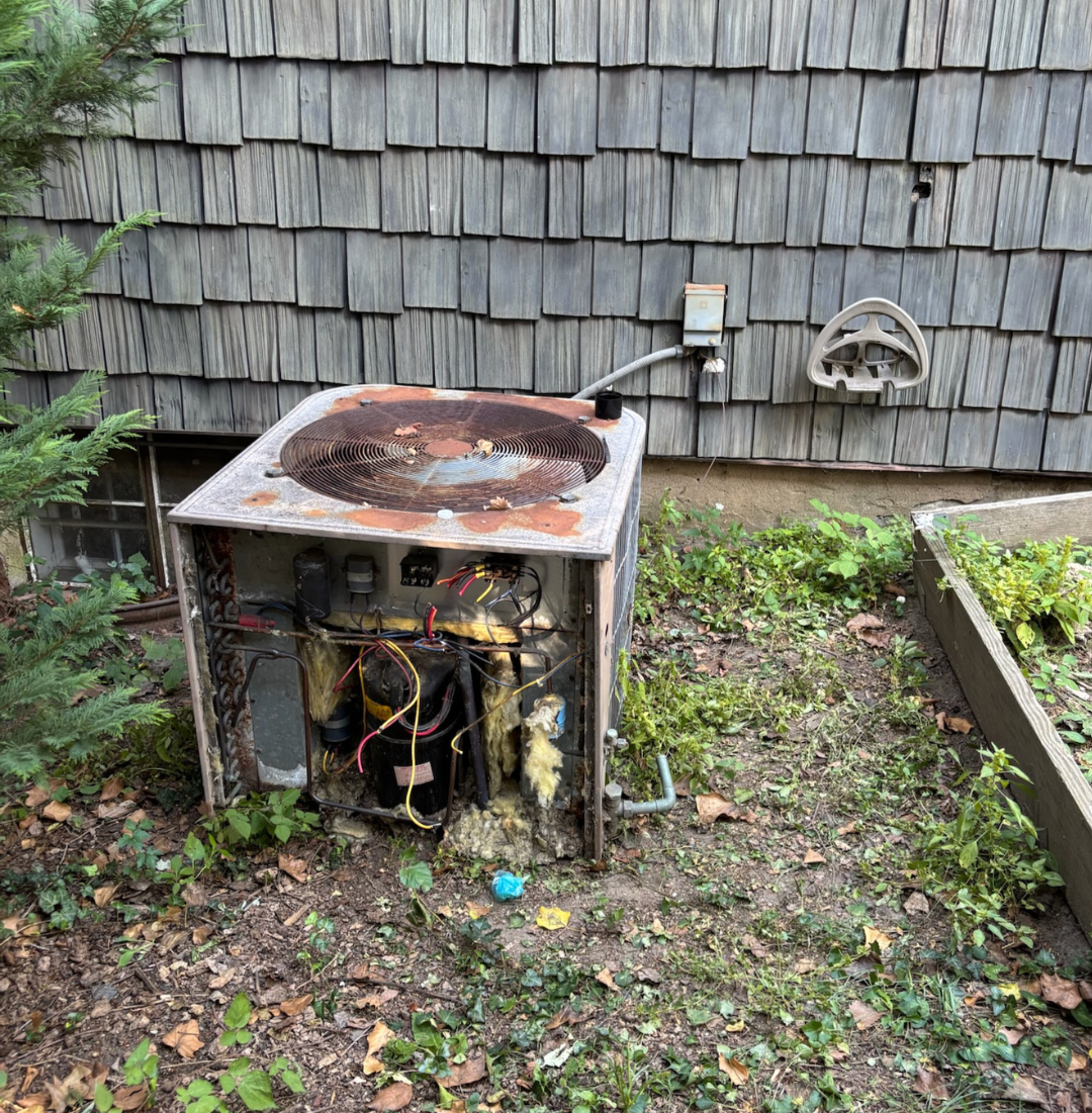 A rusty and damaged air conditioning unit with exposed components, wires, and broken parts, located outdoors near a wooden house with shingle siding and a garden bed with plants.