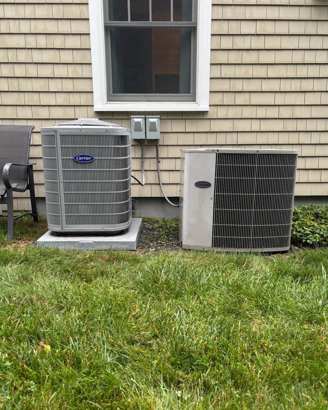 Outdoor view of two Carrier air conditioning units installed on a concrete pad outside a house with tan siding. A window and electrical boxes are visible above the units. There is green grass in the foreground.