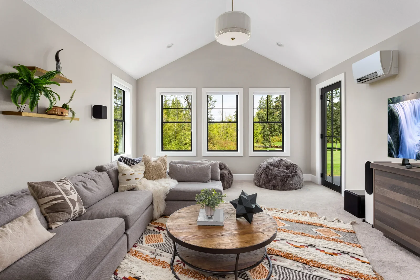 A modern living room with a large gray sectional sofa decorated with patterned and textured pillows, facing a coffee table with plants and geometric decor, in front of three large windows showing greenery outside. There's a plush pouf near the windows, a patterned rug under the table, a mounted television on a wooden console, an air conditioning unit, and a white ceiling with a hanging light fixture.