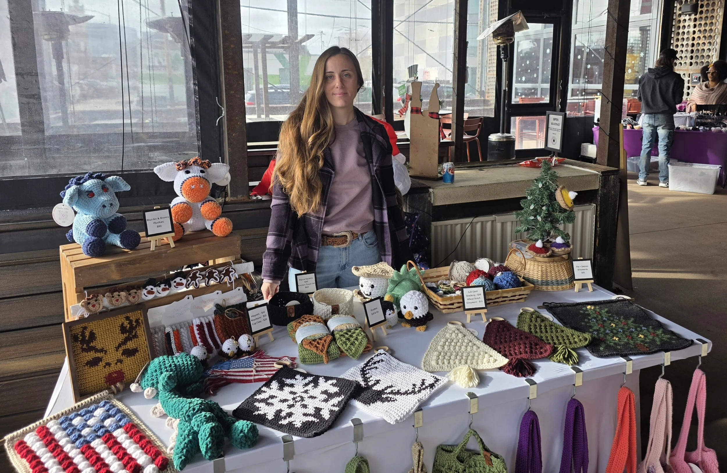 A woman stands behind a table displaying handmade crocheted crafts at a holiday market. The table features stuffed animals, Christmas-themed decorations, and knitted accessories, with a small decorated Christmas tree on the right. Other market booths and shoppers are visible in the background.