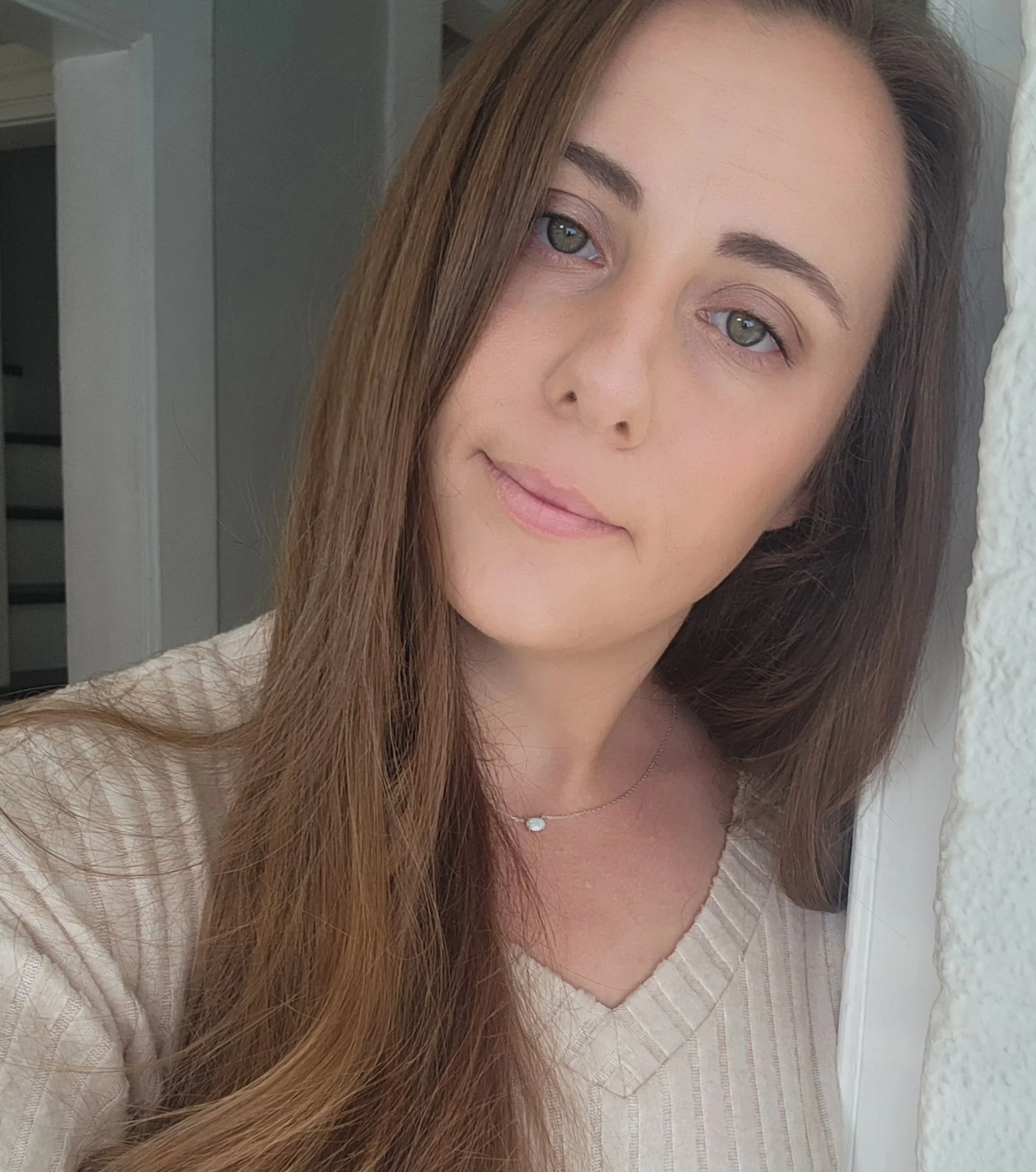 Close-up of a woman with long brown hair and blue eyes, wearing a beige top and a delicate necklace, standing near a doorway.