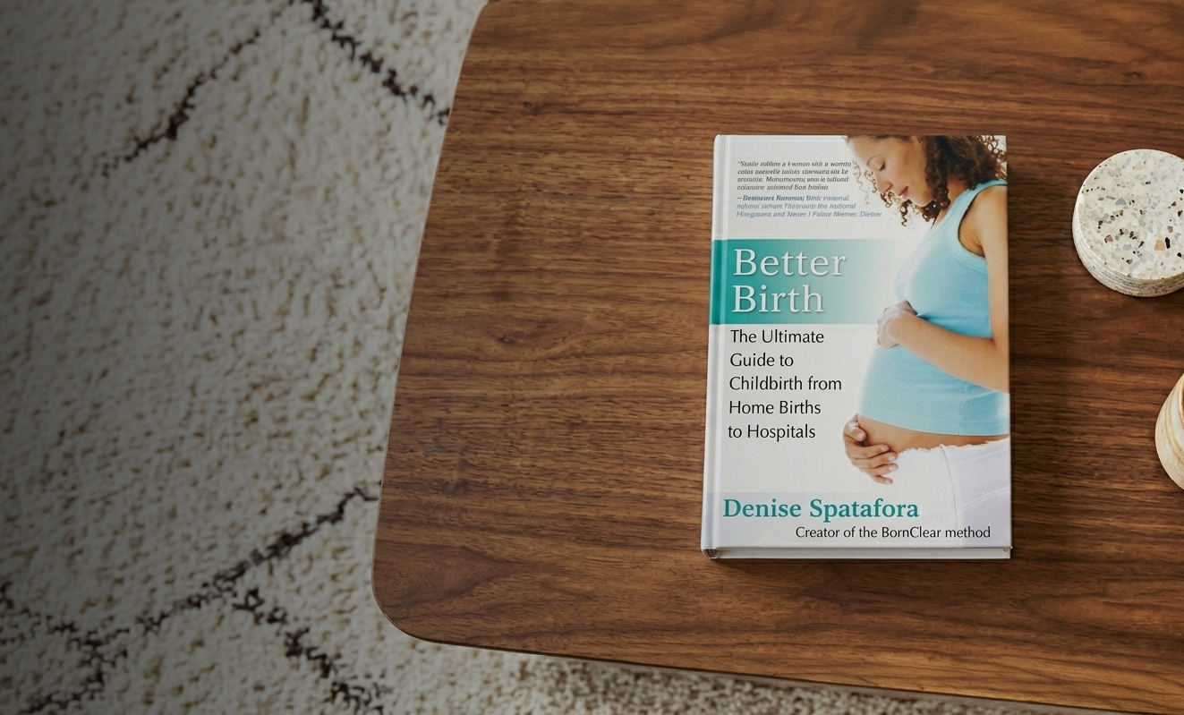 A book titled 'Better Birth' by Denise Spatafora on a wooden table, with two small decorative cups and a plant pot nearby, and a carpet partially visible on the left.