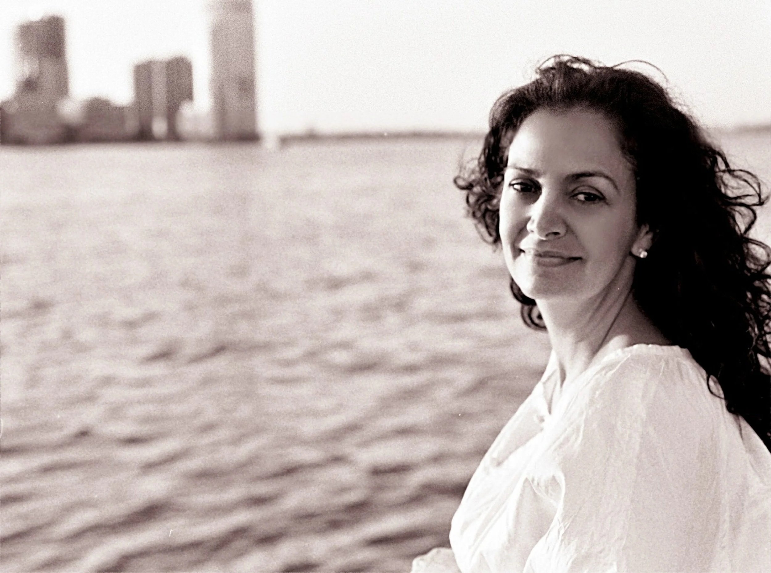 A woman with curly hair sitting by the water, looking at the camera with a slight smile, wearing a white top, with a city skyline in the background.