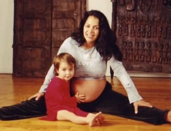 A woman with dark curly hair and a pregnant belly sitting on the floor in a split position with a young child sitting in front of her, both indoors in a room with wooden doors and floors.