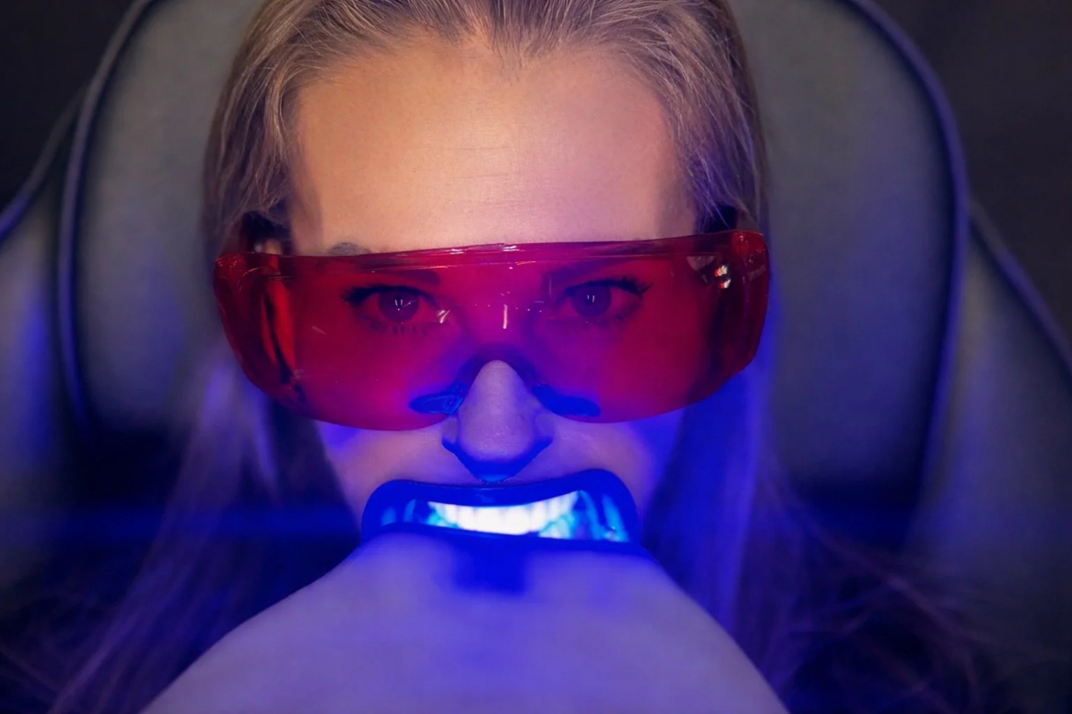 Woman smiling and holding up a shade guide for teeth color matching, with the words 'Luxe Glow' and 'Hmm' overlaid.