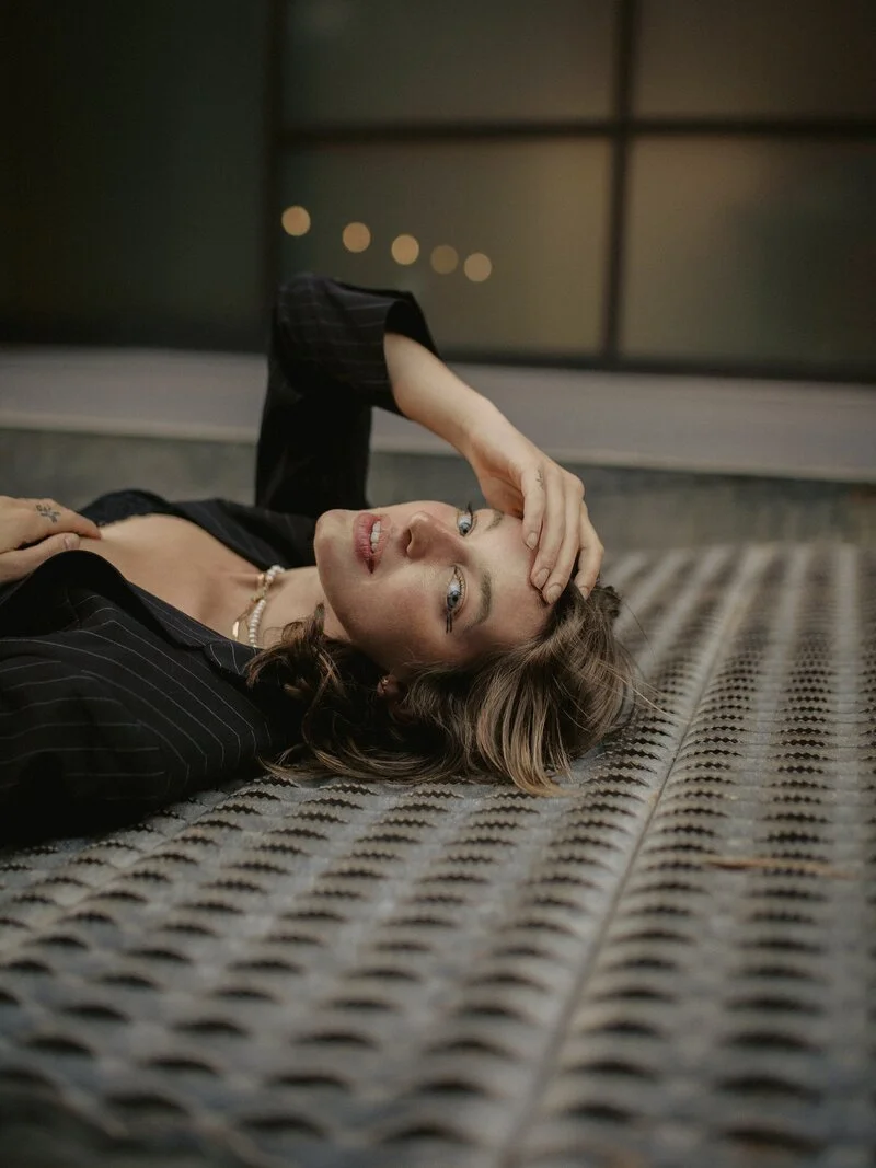 A woman with wavy brown hair lying on her back on a textured metal surface, wearing a black pinstripe blazer and pearl necklace, holding her head with her right hand, looking at the camera.