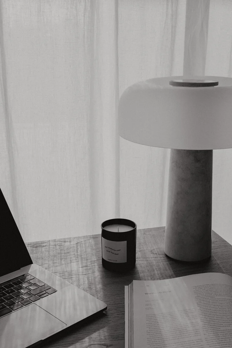 A workspace scene with an open laptop, a lit candle, a white modern lamp, and an open book on a wooden desk with sheer white curtains in the background.