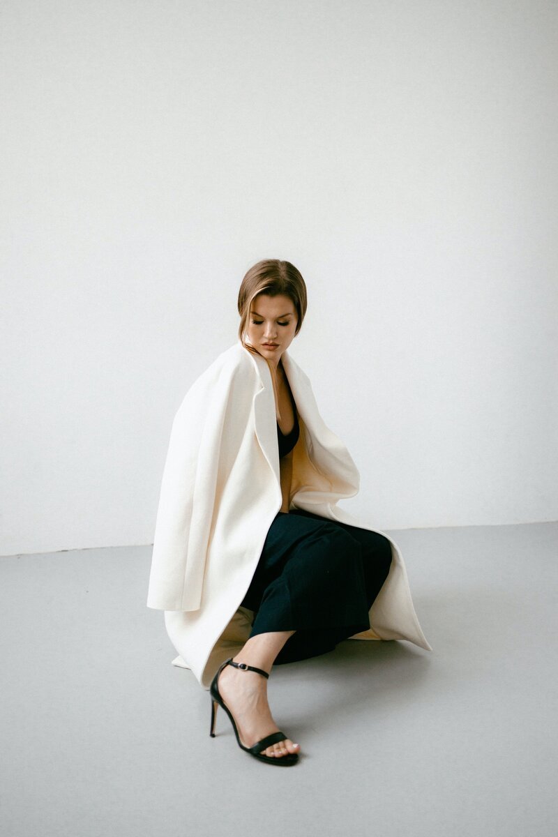 A woman with short brown hair sitting on the floor against a plain white wall. She is wearing a black dress, a white blazer draped over her shoulders, and black high-heeled sandals.
