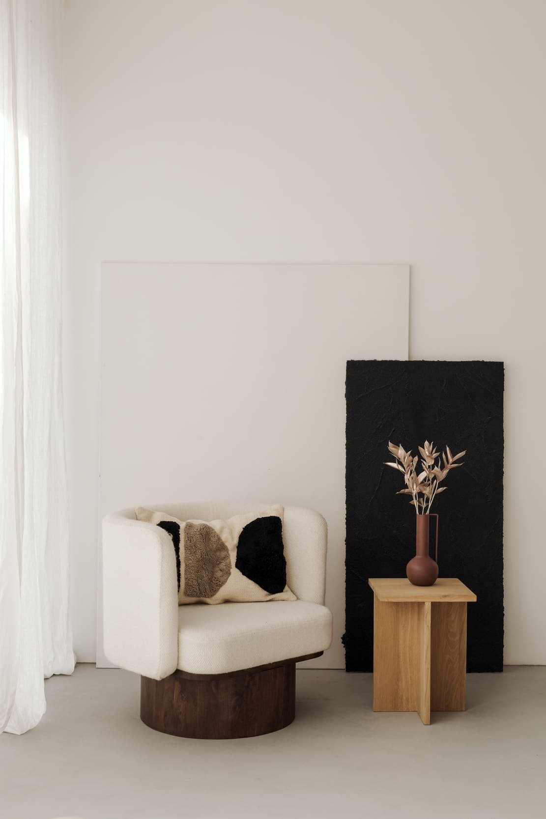 Minimalist living room corner with a white armchair, black and beige pillow, wooden side table with a pottery vase containing dried leaves, black textured panel, and white curtains.
