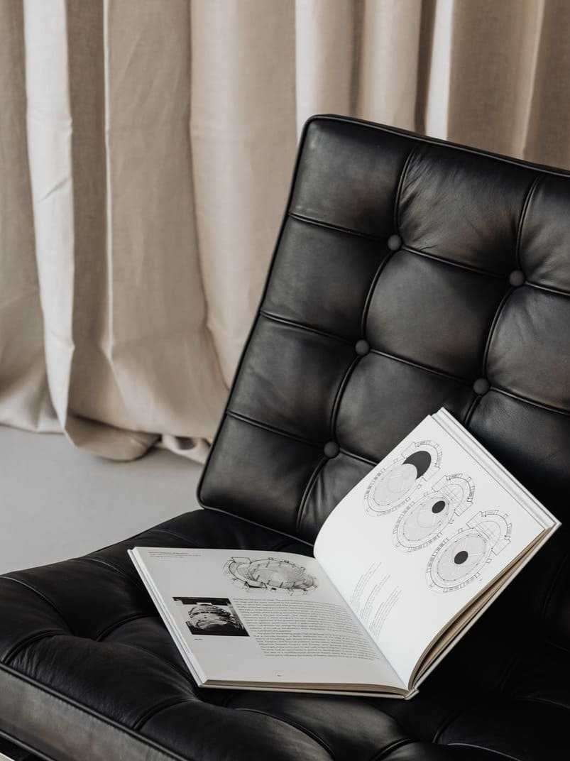 Open book with technical drawings resting on a black leather chair in front of beige curtains.