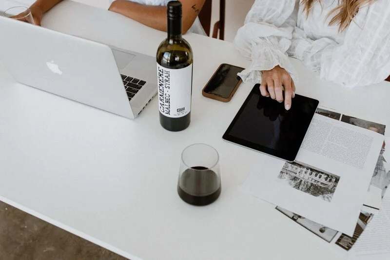 A white table with a laptop, a bottle of wine, a glass of wine, a tablet, a smartphone, and an open magazine with black-and-white photographs, with two women sitting around it.