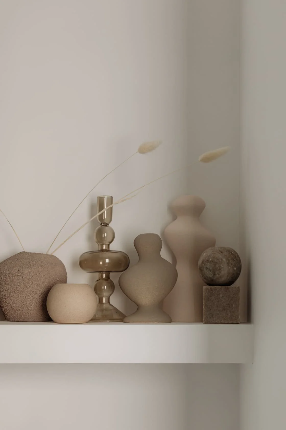Decorative neutral-colored vases and stones arranged on a white shelf, with some dried grasses placed in a transparent glass vessel.