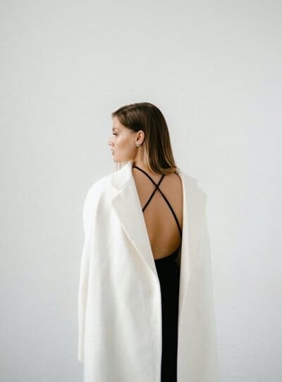 Woman wearing a black dress with strappy back, draped in a white coat, standing against a plain gray background.
