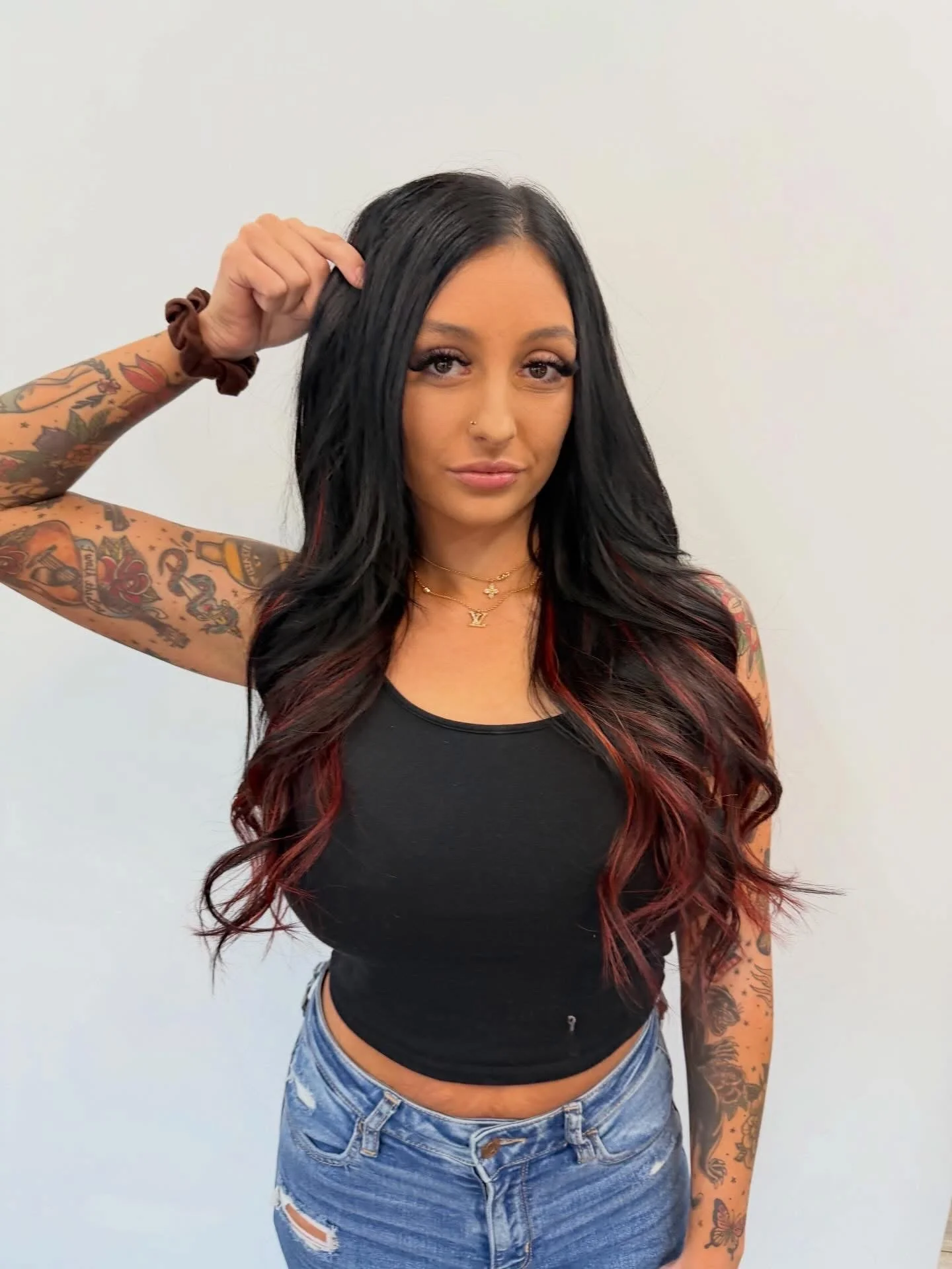 Back view of a woman with long, wavy, burgundy hair, wearing a black top and ripped jeans, inside a salon or similar setting.