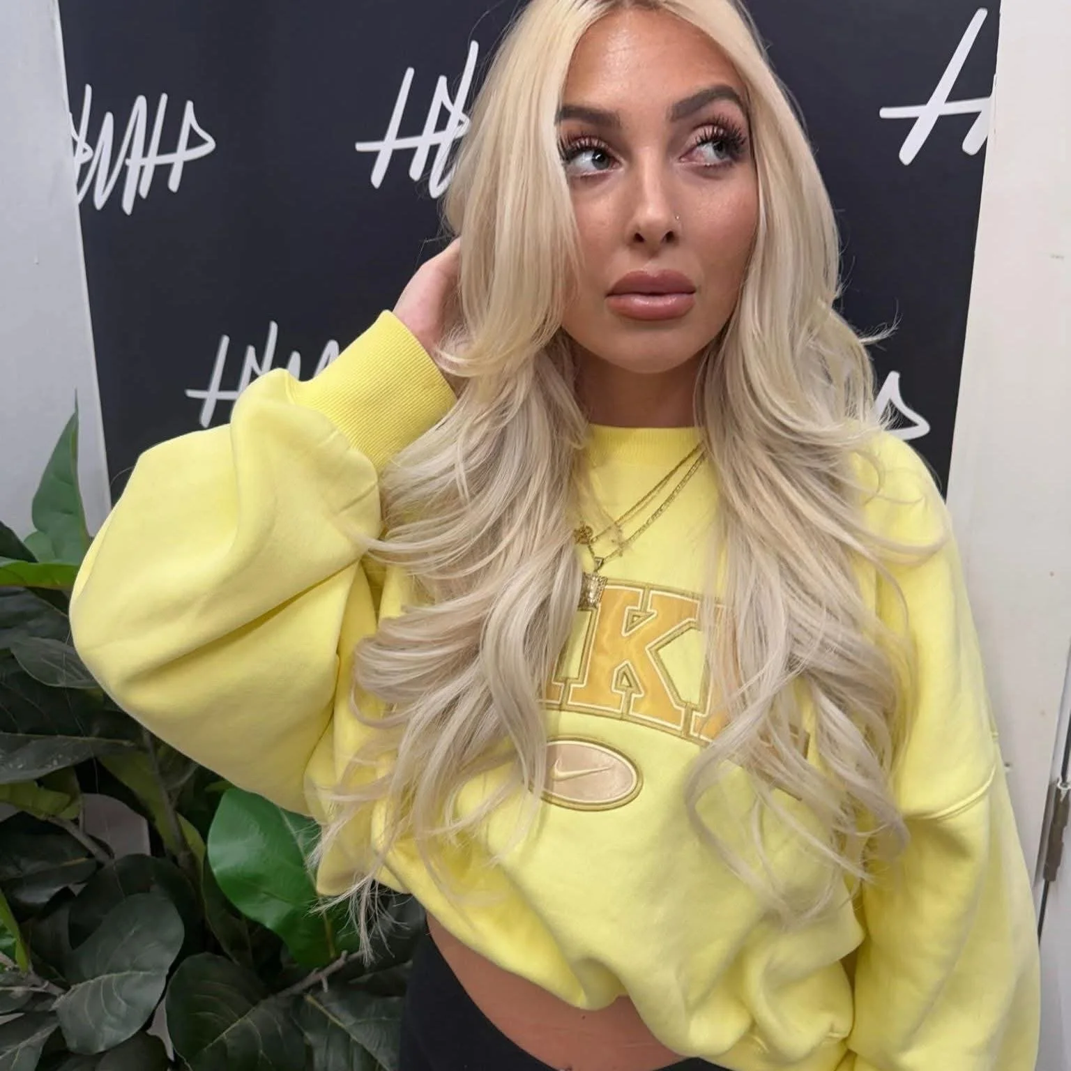 A woman with long, wavy blonde hair, wearing a black top with a striped neckline, a silver necklace, and a silver ring on her finger, standing indoors.