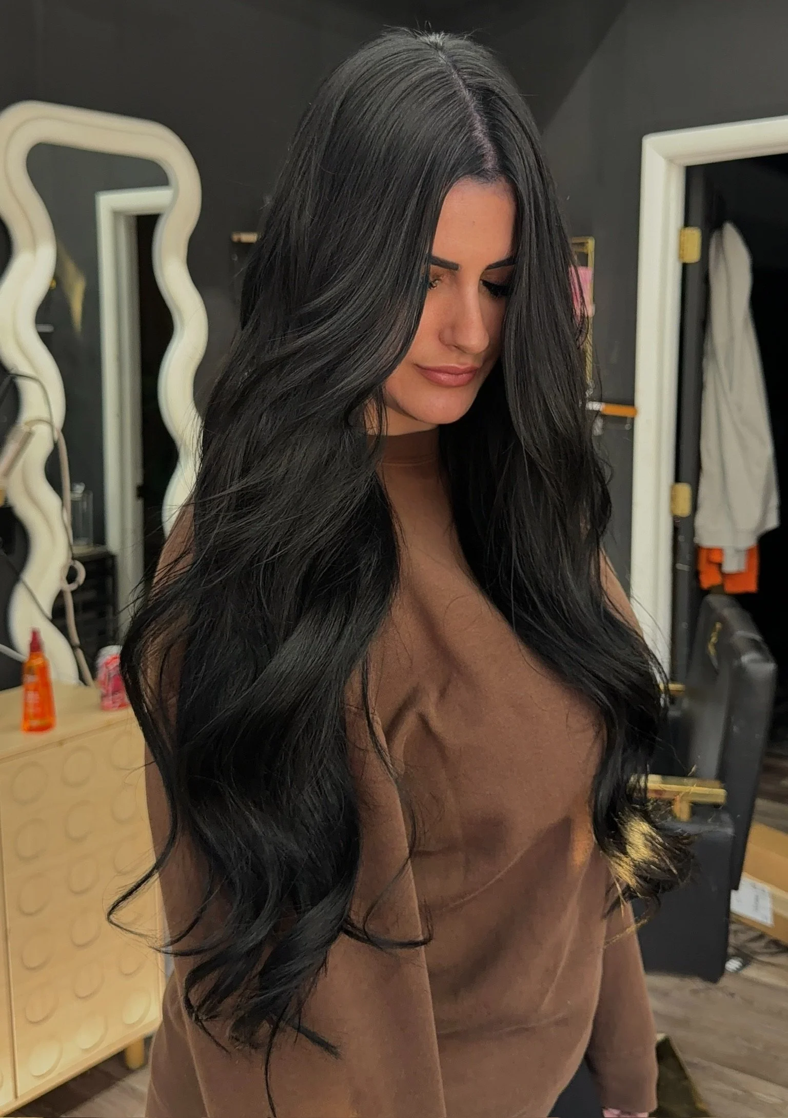 Back view of a woman with long, wavy, burgundy hair, wearing a black top and ripped jeans, inside a salon or similar setting.