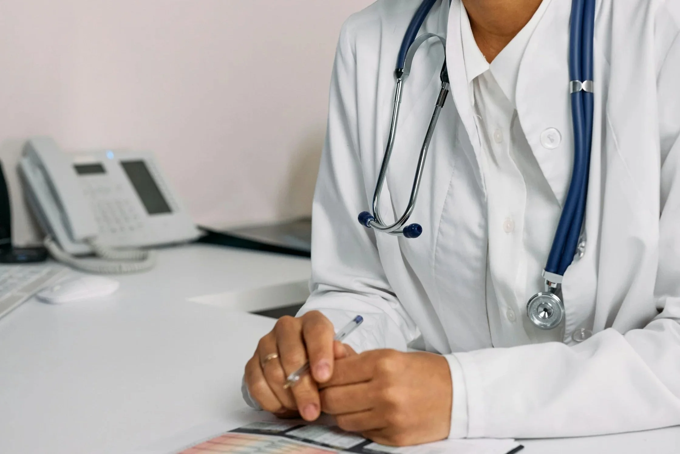 A healthcare professional wearing a white coat and a stethoscope around their neck, sitting at a desk and writing on a piece of paper with a pen.