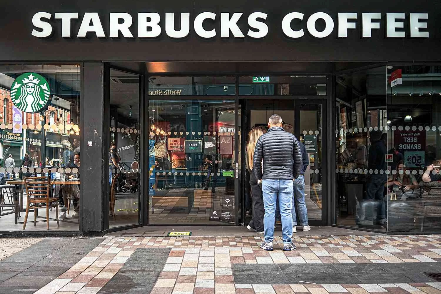 Exterior view of a Starbucks coffee shop with customers entering and inside. The storefront has large glass windows with the Starbucks logo and black framing, and a sign above that reads "STARBUCKS COFFEE" in large white letters.