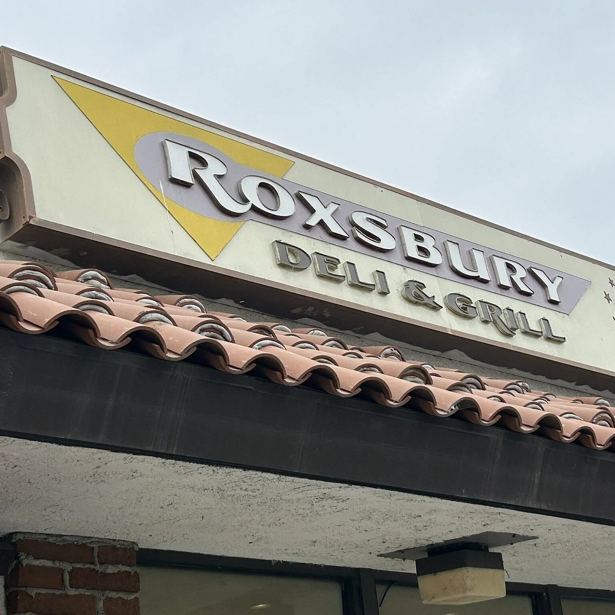 Signboard for Roxbury Deli & Grill above a tiled roof and brick wall