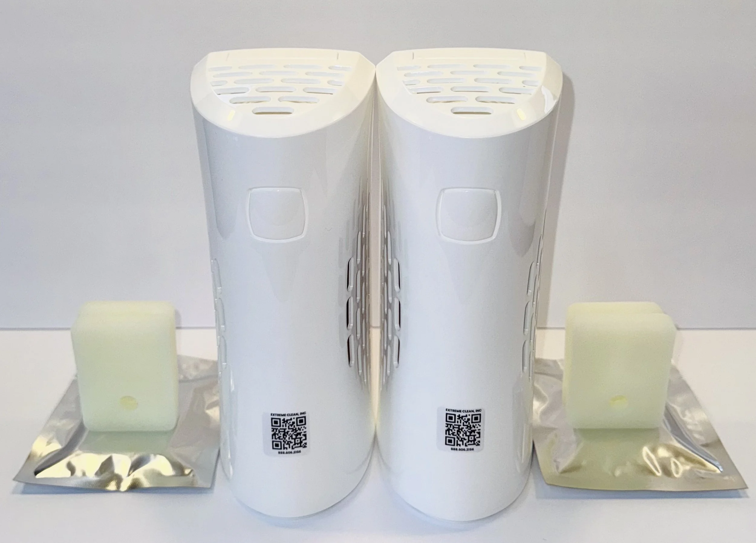Two white cylindrical air purifiers with vents on top, placed side by side on a surface, with two packs of green foam filters in front of each.