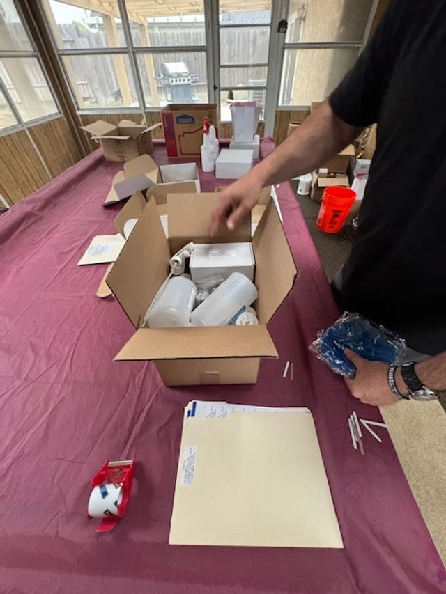 Vince sorting plastic cups inside an open cardboard box on a table covered with a maroon tablecloth, with various supplies and paper towels in the background.