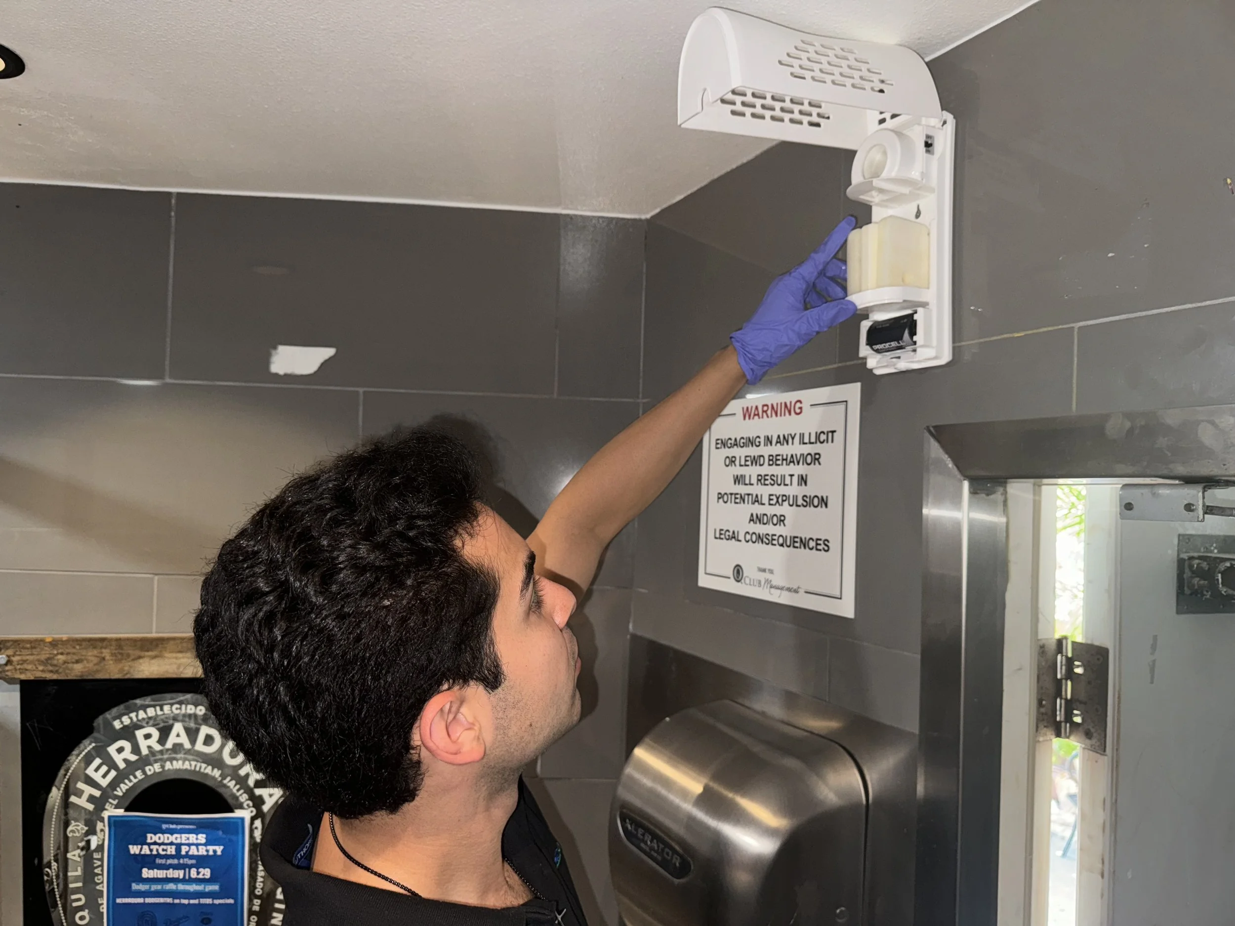 Vince wearing a purple glove reaches up to adjust or inspect a fire alarm device on a gray tiled wall, with a warning sign below.
