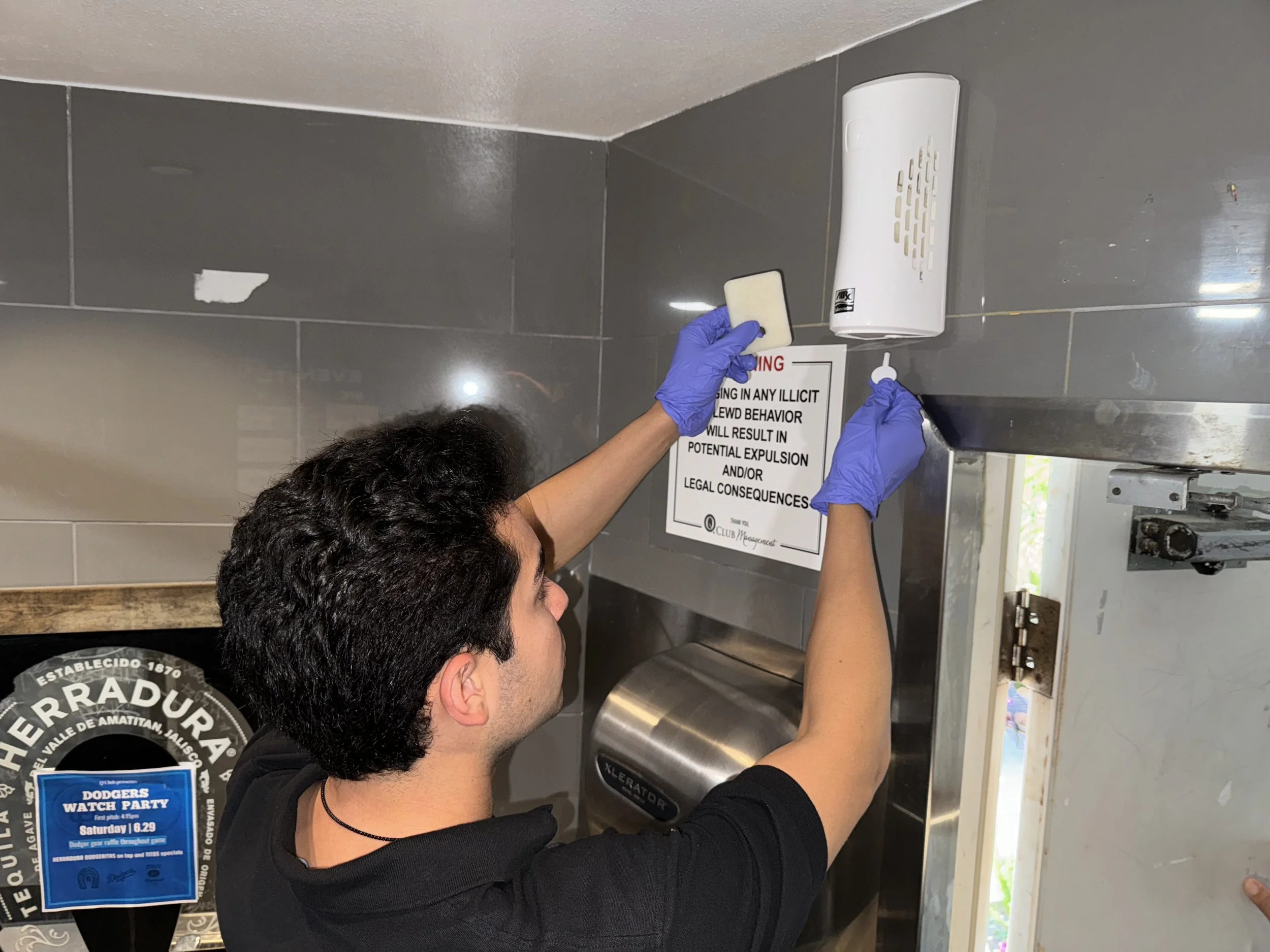 Vince in a black ROC-D shirt wearing blue gloves is cleaning or refilling a hand sanitizer dispenser on a tiled wall. He is holding a sponge in one hand and a tool in the other. There is a safety sign on the wall above the dispenser.
