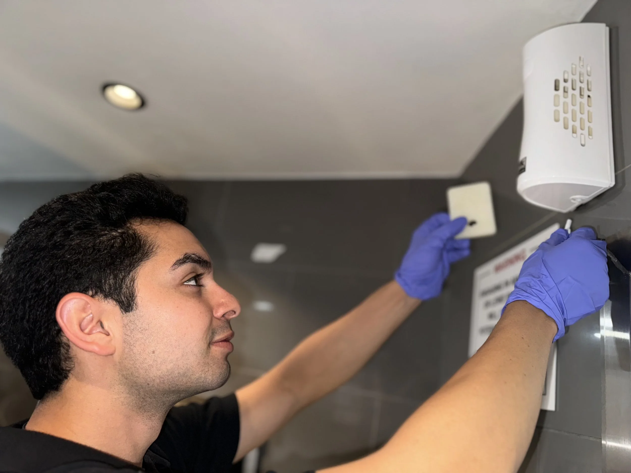 Vince wearing blue gloves installing a white device on a kitchen or bathroom wall.