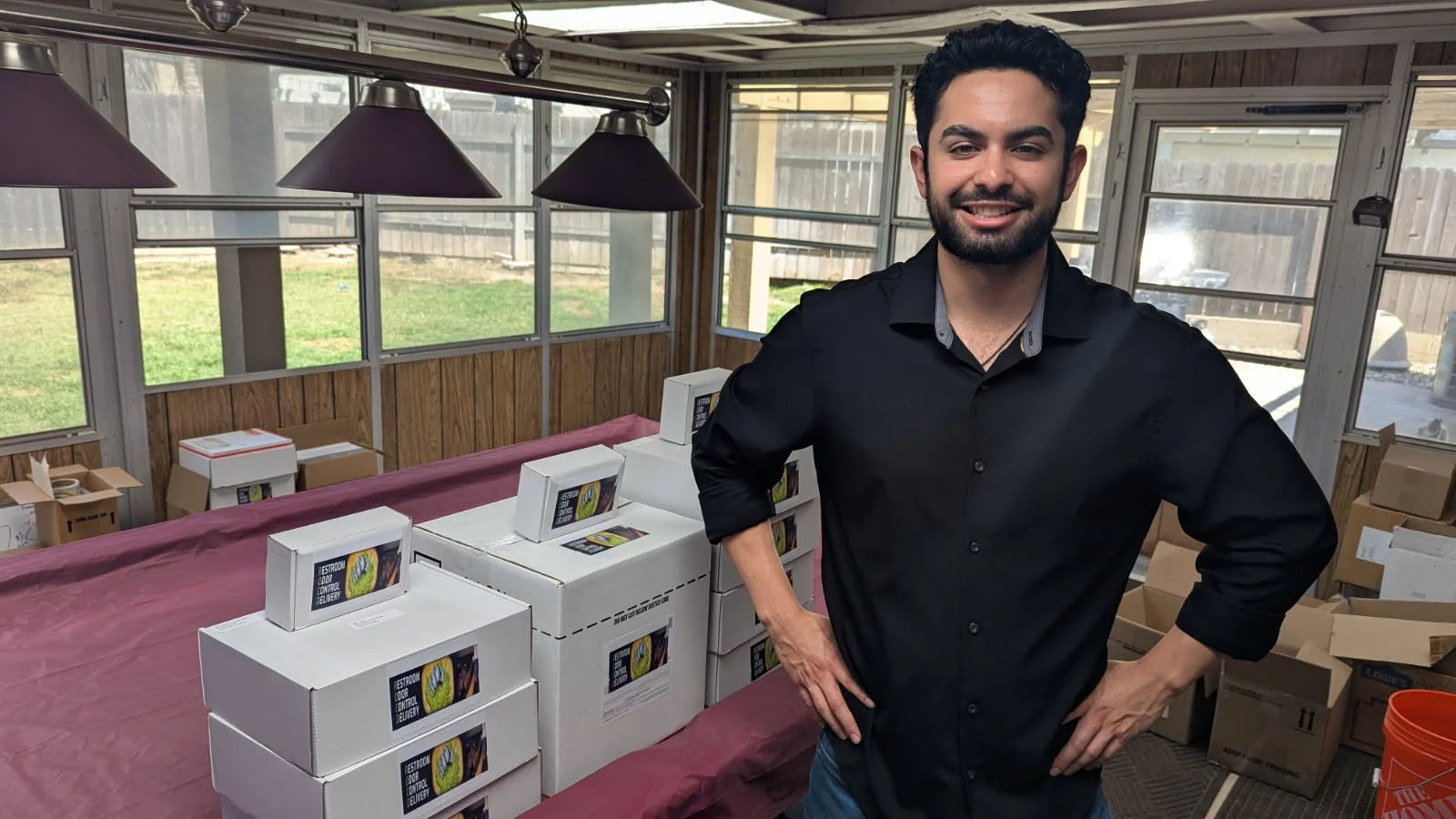 Vince, smiling, wearing a black shirt, standing in a room with boxes and packages on tables, with large windows and wooden walls in the background.