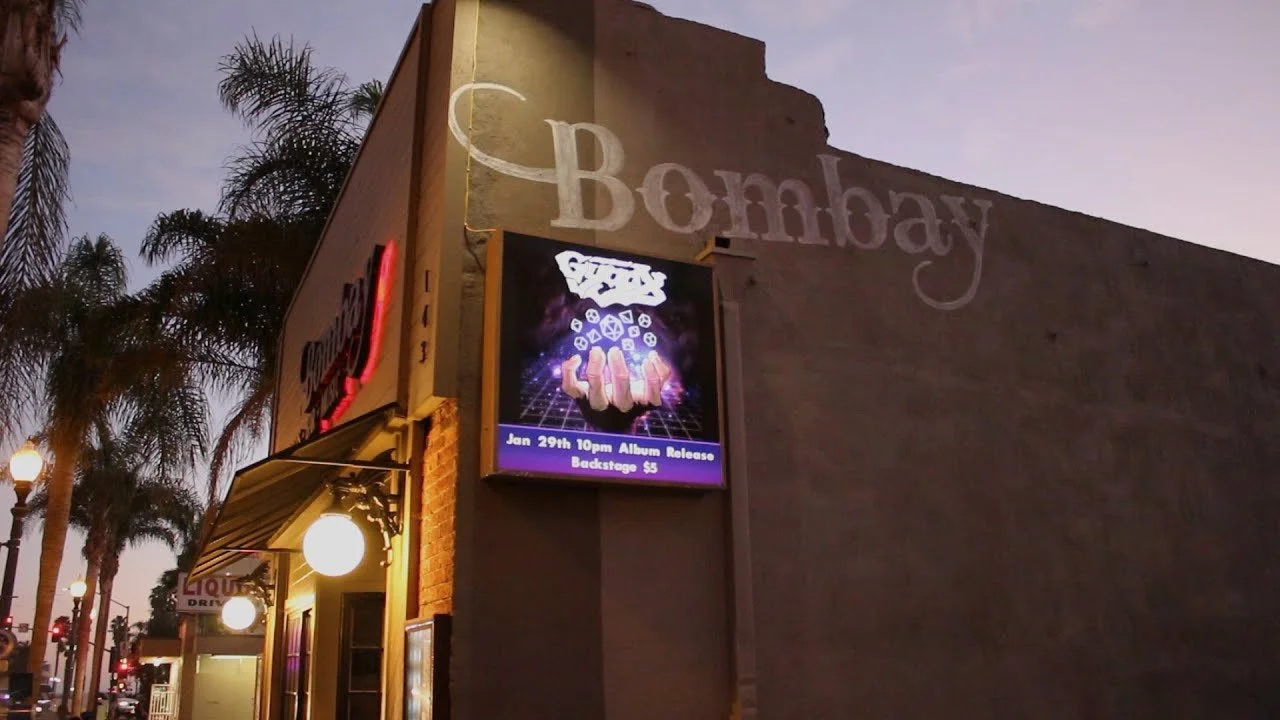 The exterior of a nightclub or bar called Bombay, with a digital sign advertising an album release event on January 29th at 10 pm, with backstage tickets costing $5. The building is illuminated with streetlights, and palm trees are visible in the background during dusk.