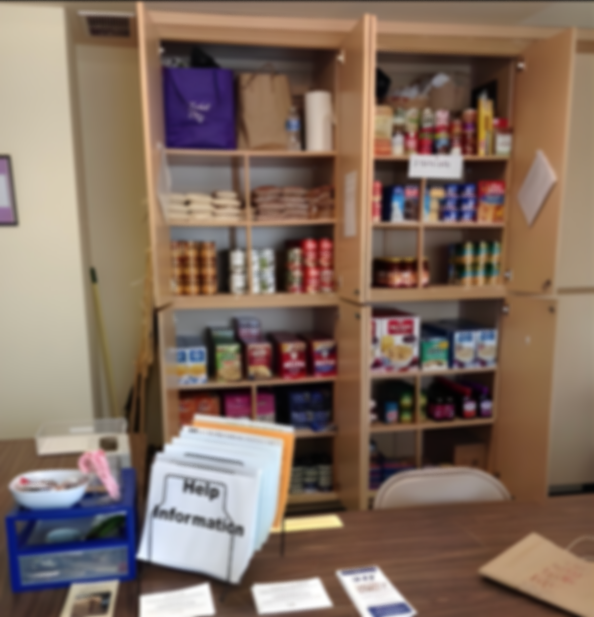 Blurred image of a wooden cabinet filled with food items and canned goods, with a table in front holding informational brochures and a snack bowl.