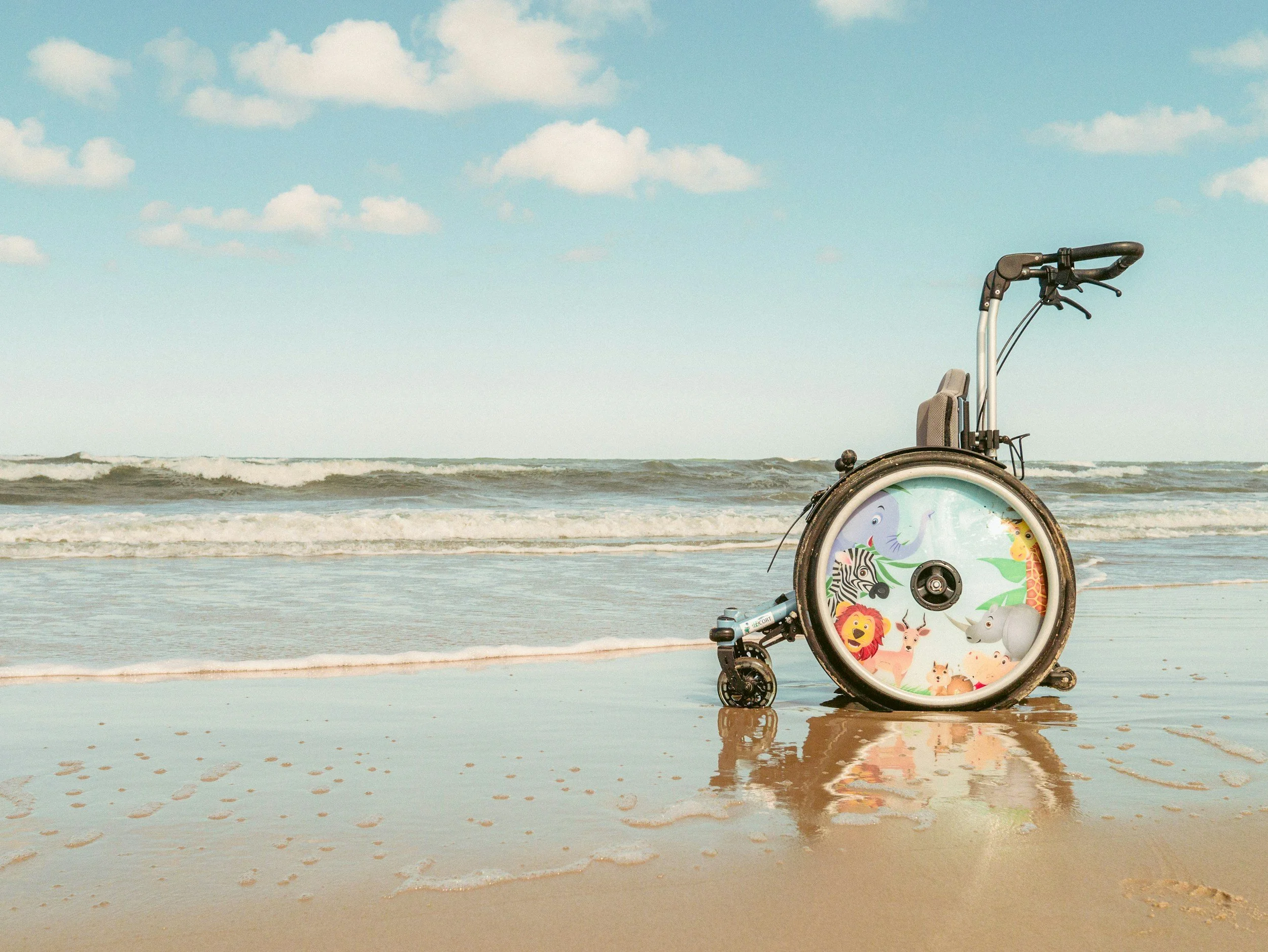 A wheelchair with animal illustrations on the wheel cover is positioned on a wet sandy beach near the ocean with small waves, under a partly cloudy sky.