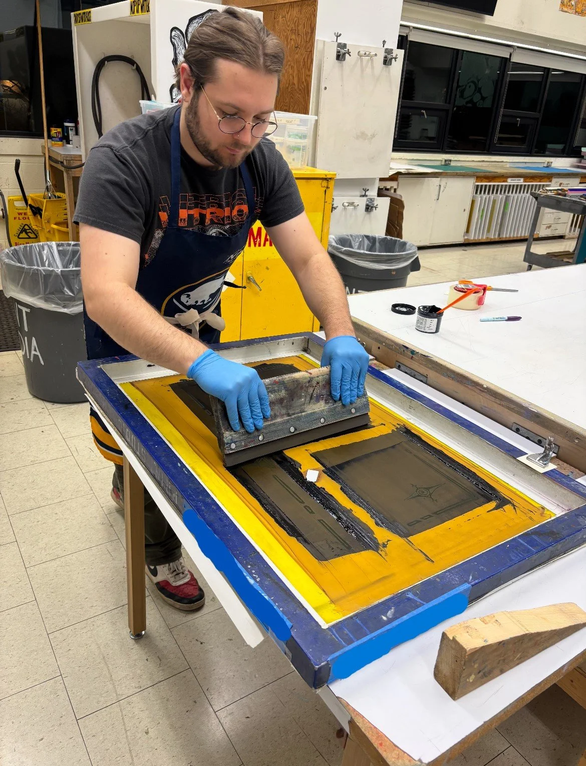 The artist, a man wearing glasses, a black t-shirt, an apron, and blue gloves is working with a large printmaking screen on a table in an art studio or workshop.