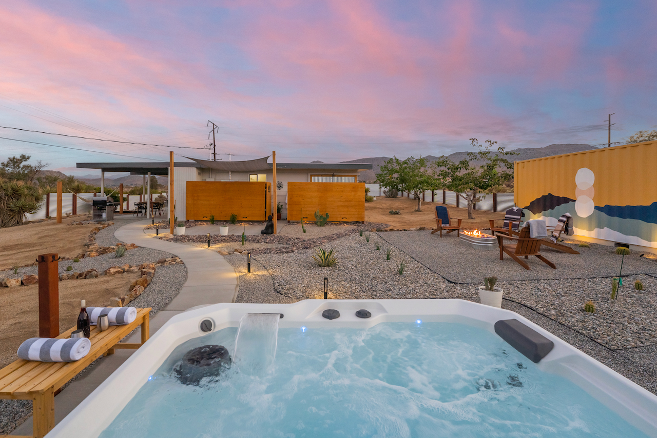 A backyard with a hot tub, a pathway, lounge chairs around a fire pit, desert plants, and a house with a covered patio during sunset.