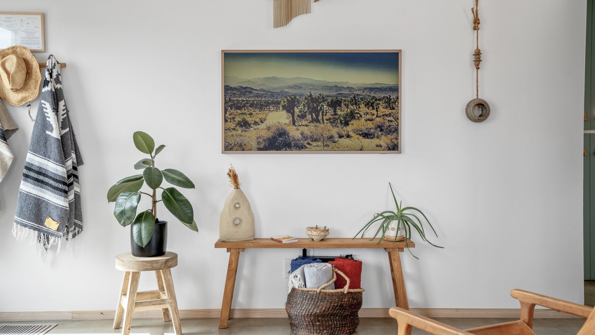 Living room wall with a framed desert landscape photograph, hanging rope decoration, a small side table with a potted rubber plant, natural fiber vase, and a decorative bowl, and a woven basket with blankets on the floor.
