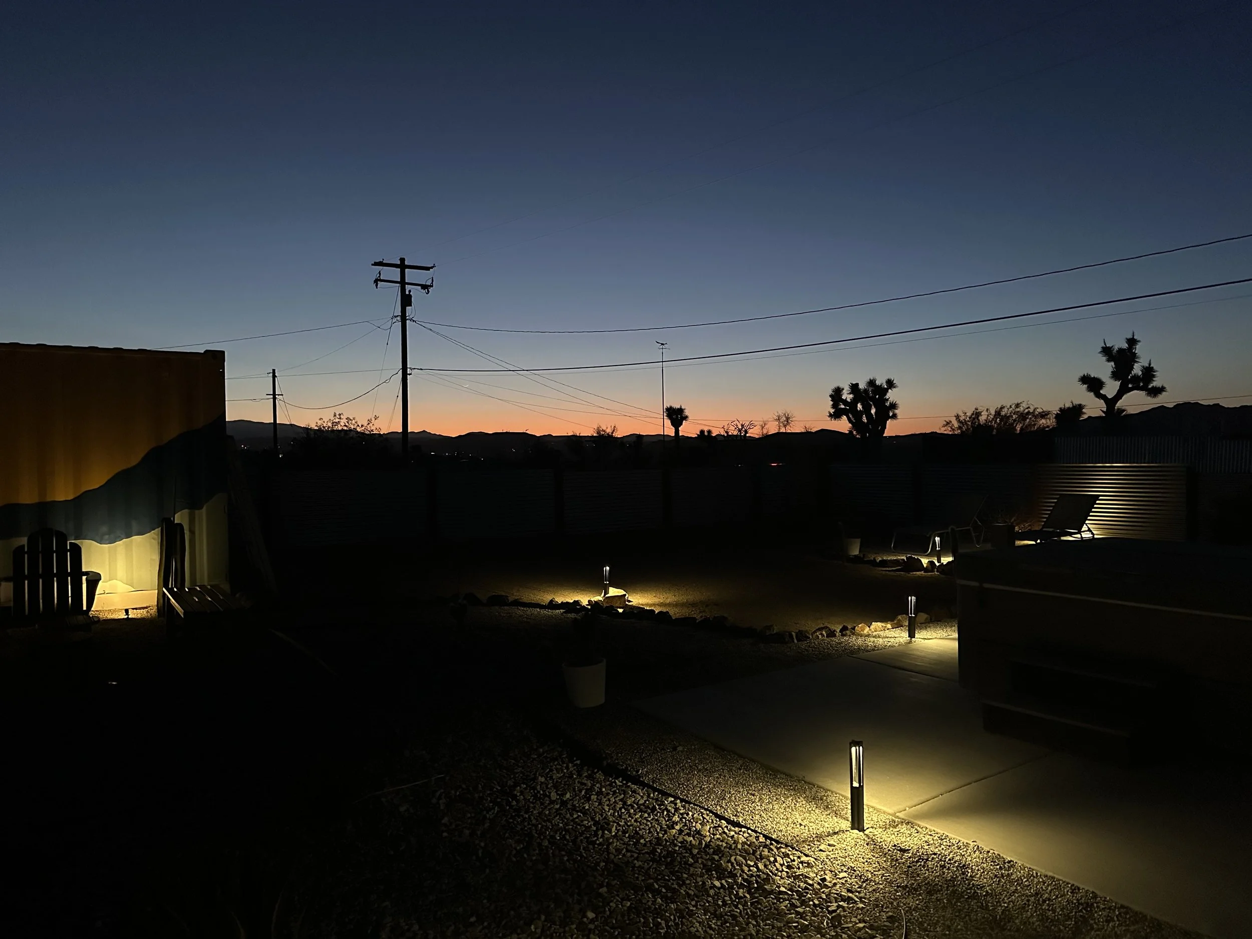 A backyard at dusk with three illuminated pathway lights, chairs, and desert trees silhouette against a twilight sky with a faint orange glow near the horizon.