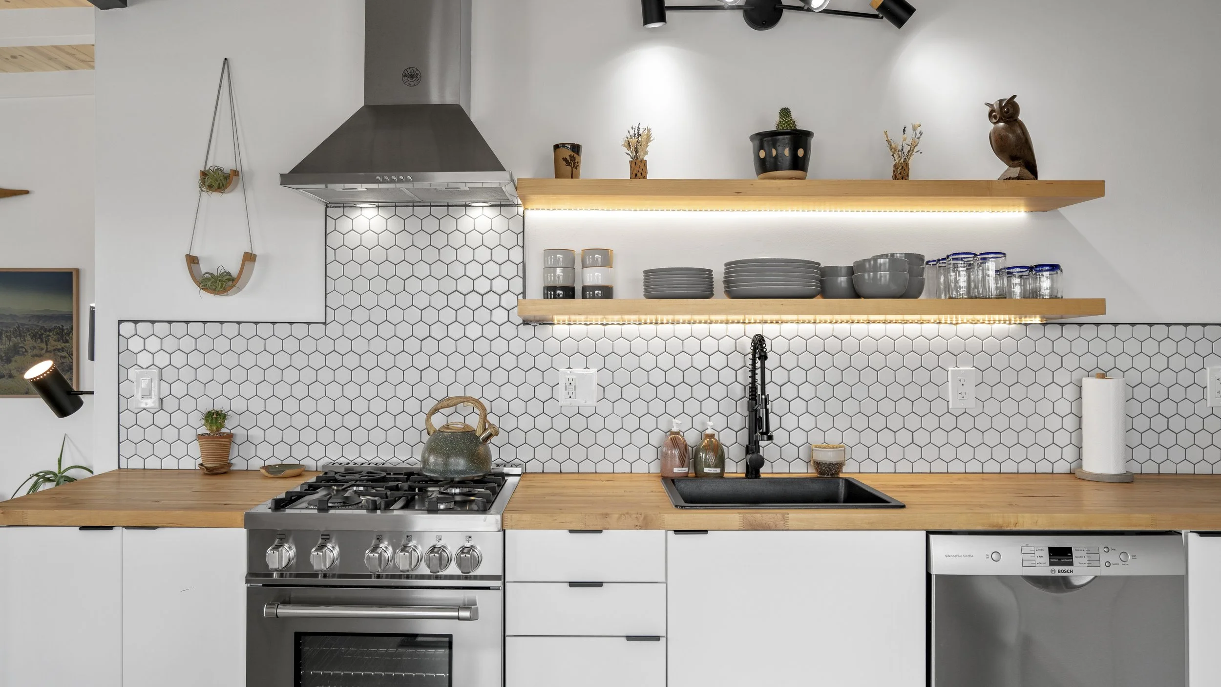 Modern kitchen with white cabinets, wooden countertops, stainless steel oven, black sink, hexagonal tile backsplash, open wooden shelves with dishes, glass jars, decorative owl, and plants.