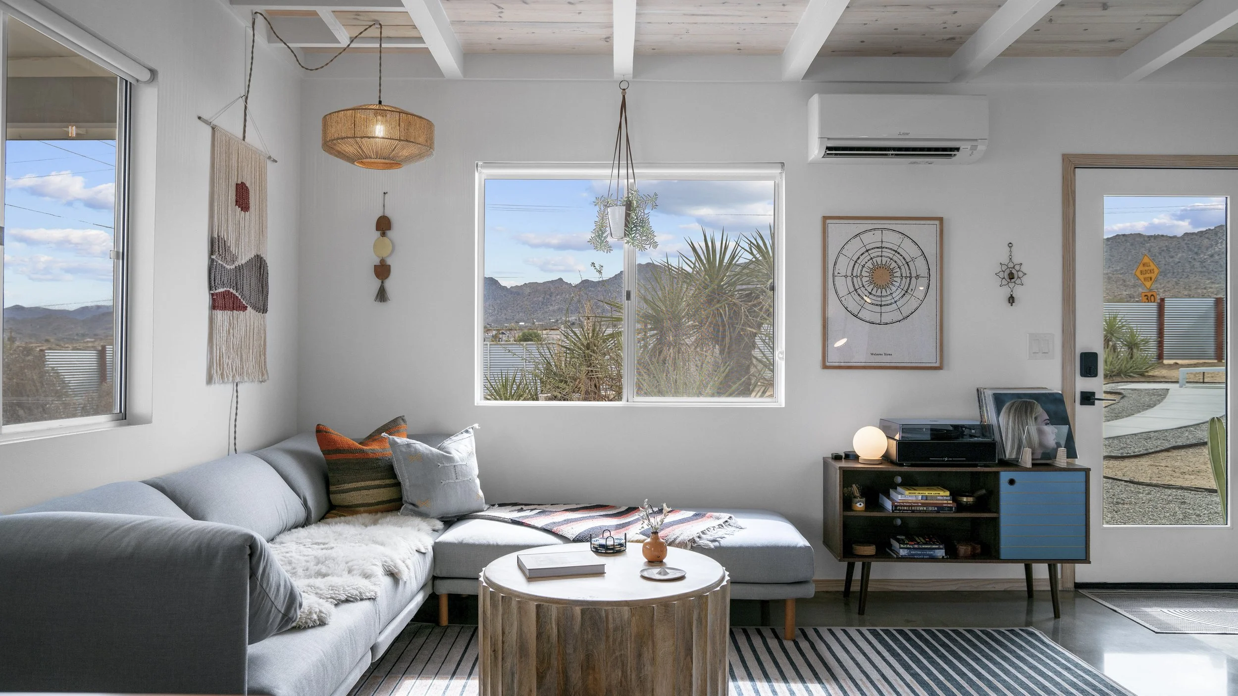 Living room with white sectional sofa, colorful pillows, wooden coffee table, and a blue cabinet holding a record player and framed photo. Decor includes wall hangings, a hanging light, and a view of desert mountains outside windows.