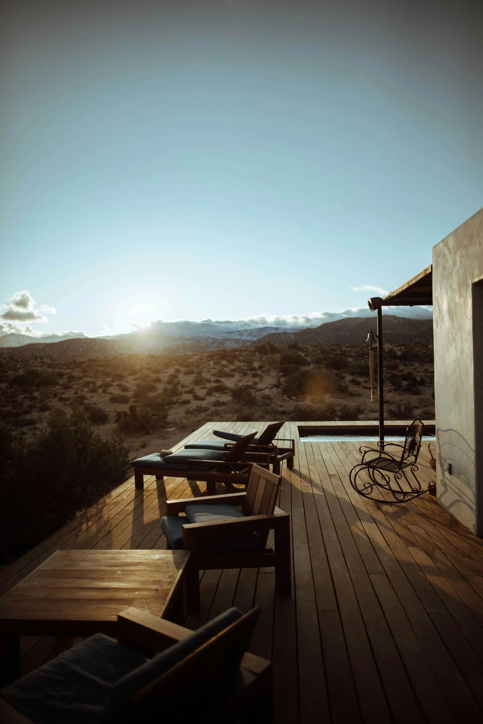 Sunset view from a wooden deck with outdoor furniture, including chairs and lounge chairs, overlooking a desert landscape with mountains in the distance.