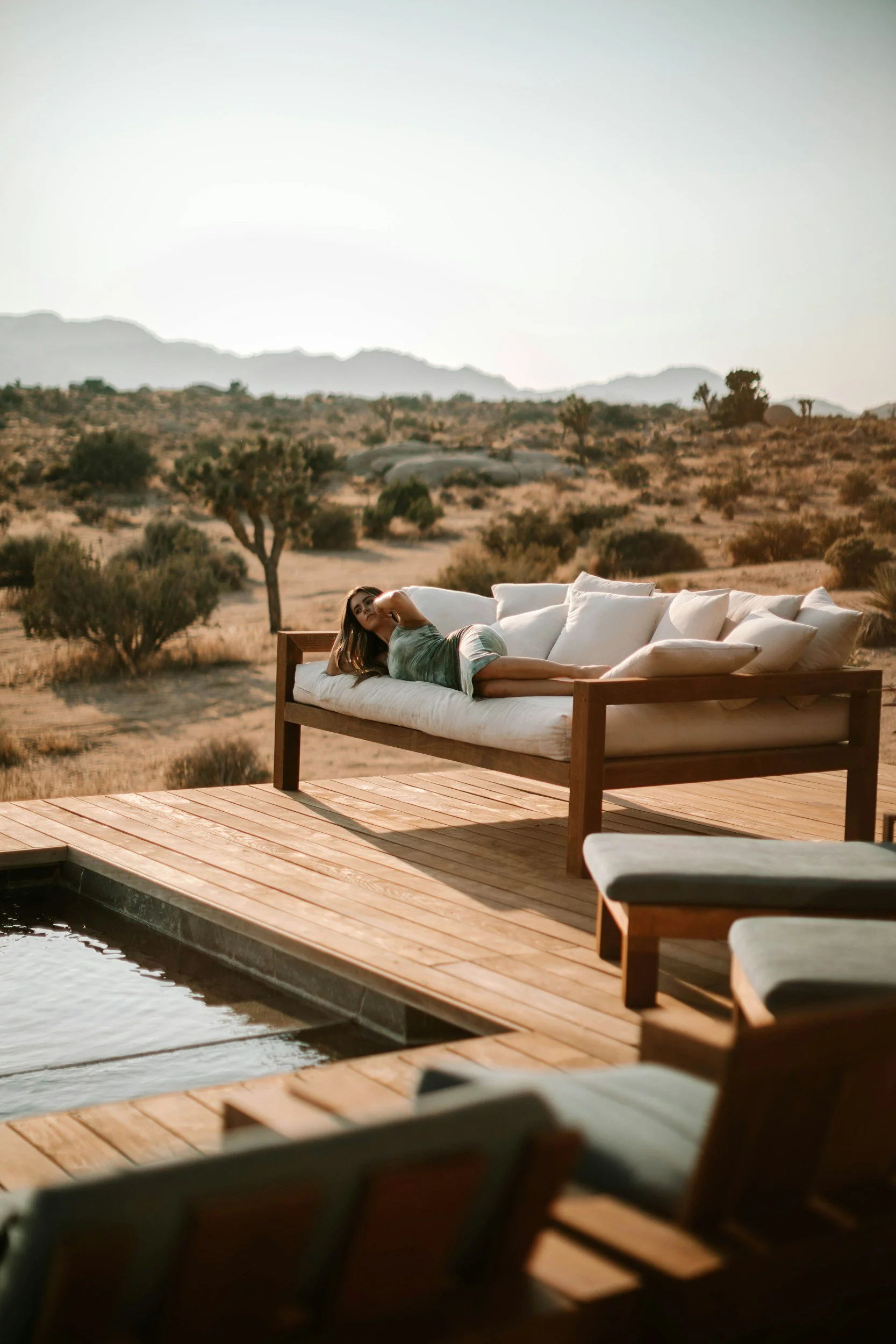 A woman relaxing on an outdoor daybed on a wooden deck in a desert landscape at sunset.
