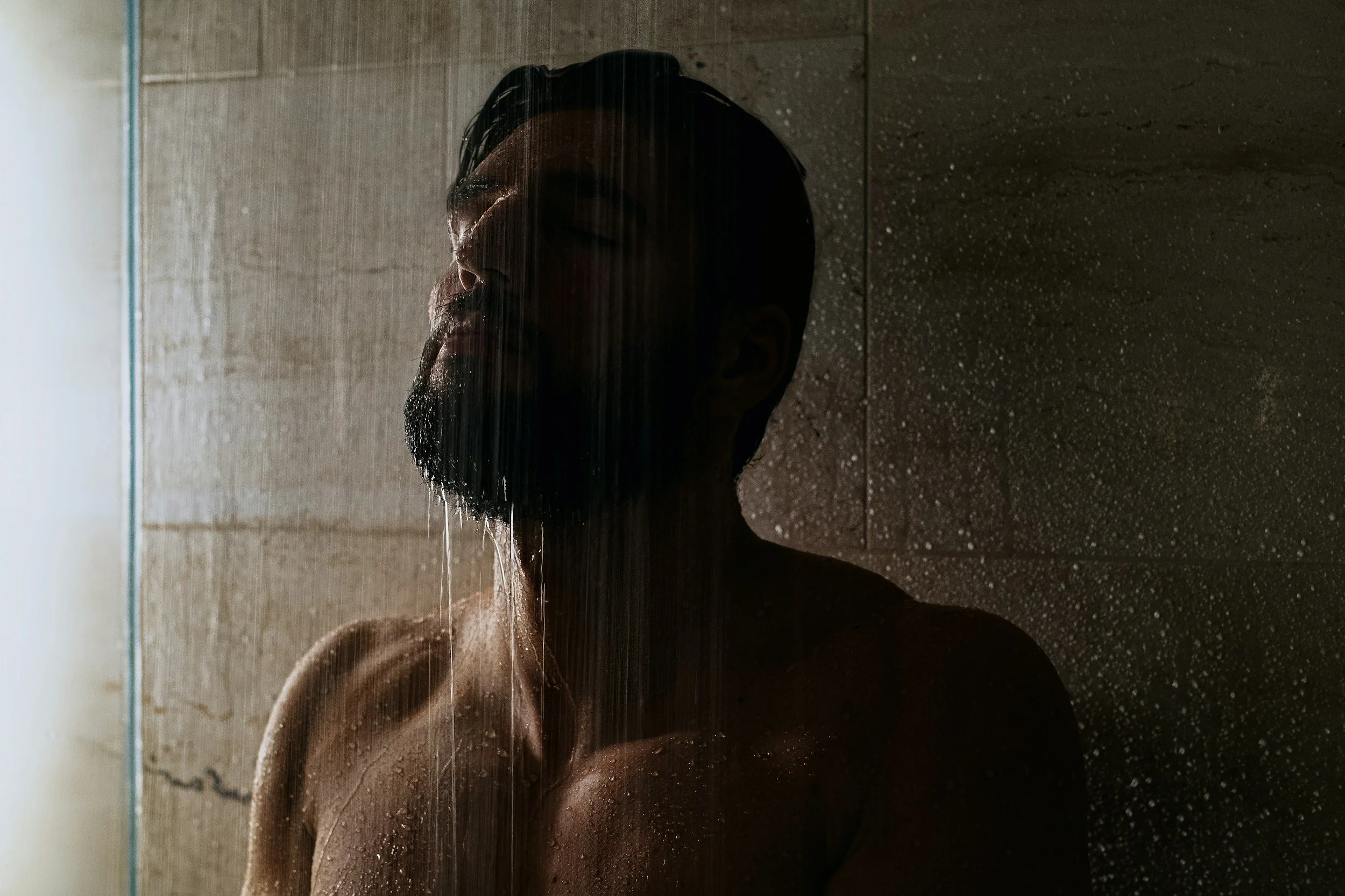 A man with a beard taking a shower, with water streaming down on his face and shoulders, in a dimly lit bathroom.