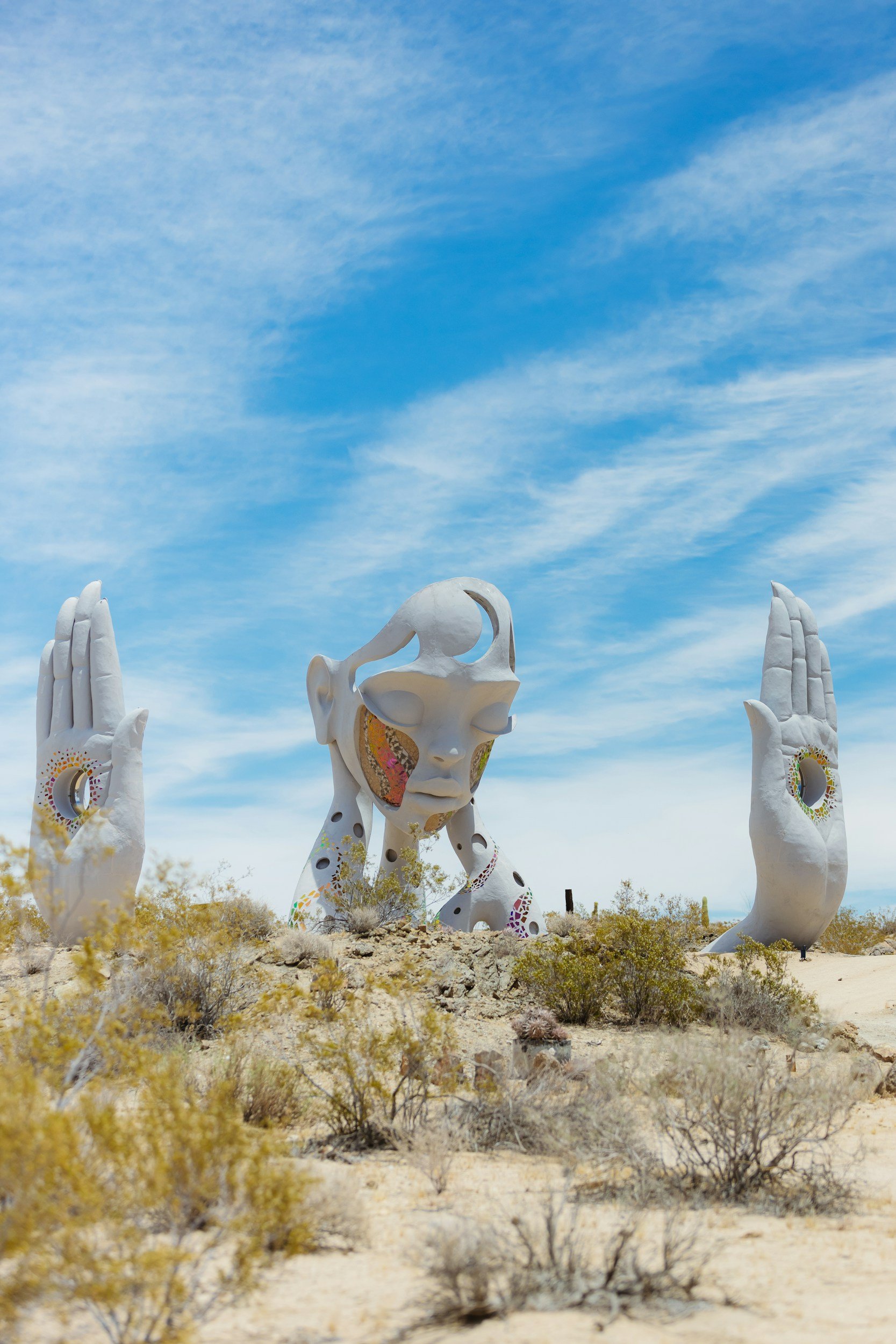 Large outdoor sculpture of a human face and hands with closed eyes, in a desert landscape with sparse bushes and a blue sky with clouds.