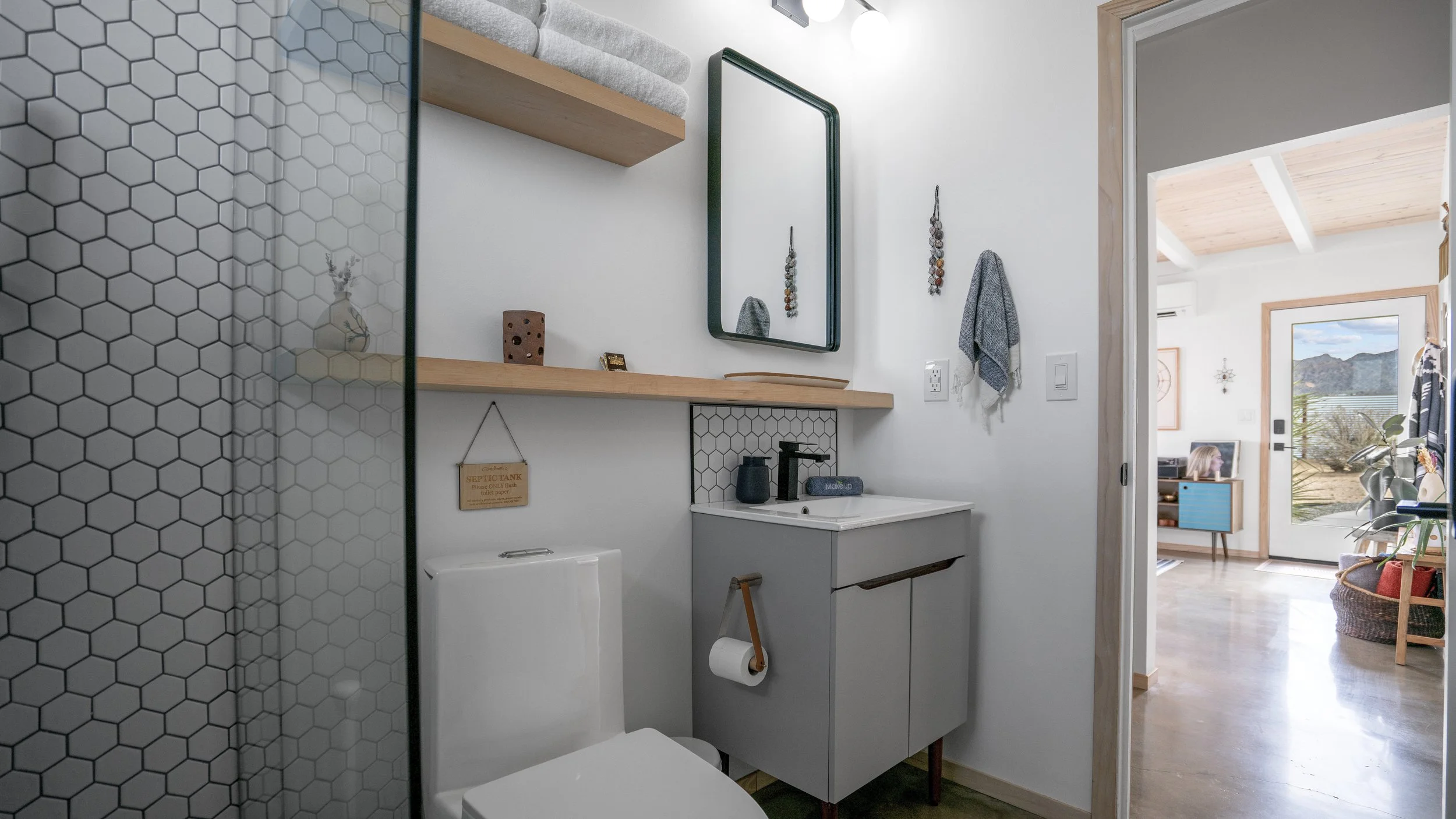 Small modern bathroom with a toilet, small sink with black faucet, mirror above, wood shelf with towels, and hexagon tile wall in shower. View into a bright living room through door.